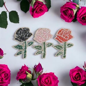 Könnte beinhalten: Drei juwelenbesetzte Rosen-Patches in Schwarz, Rosa und Rot. Jede Rose ist mit Strasssteinen verziert und hat einen grünen Stiel und Blätter.