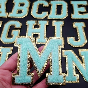 May include: A set of turquoise blue felt letters with gold glitter trim. The letters are in the style of varsity or collegiate letters. The letters are in the alphabet from A to N.