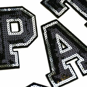 Peut inclure: Patchs thermocollants en sequins noirs et blancs en forme de lettres P, A et Y. Les lettres ont un contour blanc et sont faites de petits sequins ronds.