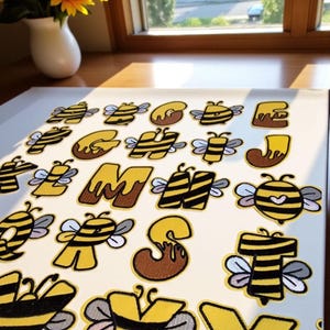 May include: Embroidered patches in the shape of letters, each featuring a bee design with yellow and black stripes and wings. The patches are on a white surface. A vase of sunflowers is visible in the background.