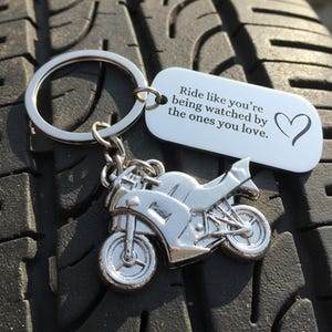 Pode incluir: Um porta-chaves de motocicleta prateado com uma etiqueta a condizer. A etiqueta tem o texto "Ride like you're being watched by the ones you love." e um símbolo de coração. O porta-chaves está num pneu preto.