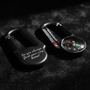 Op de afbeelding: Twee zwarte karabijnhaken. E&eacute;n is voorzien van de gegraveerde tekst 'So you always find your way home!' en de andere bevat een ge&iuml;ntegreerd rond kompas met een rode naald en een kleine thermometer met een rode vloeistofindicator.