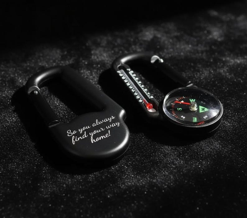Op de afbeelding: Twee zwarte karabijnhaken. E&eacute;n is voorzien van de gegraveerde tekst 'So you always find your way home!' en de andere bevat een ge&iuml;ntegreerd rond kompas met een rode naald en een kleine thermometer met een rode vloeistofindicator.