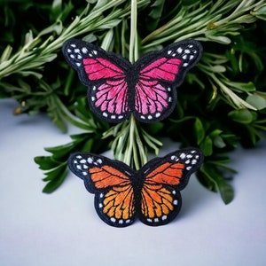 May include: Two embroidered butterfly patches, one pink and one orange, with black outlines and white accents. The patches are on a white background.