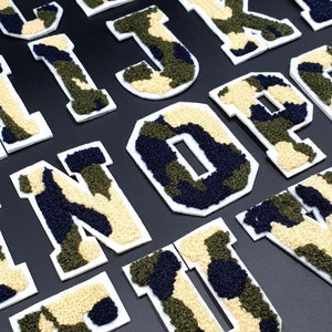 May include: A set of felt letters in a camouflage pattern. The letters are in uppercase and are all white with a black outline. The letters are arranged in alphabetical order.