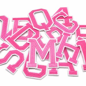 May include: A collection of hot pink embroidered varsity-style letter patches with a thick white border. The letters are arranged in a scattered pile, featuring various characters including M, Q, B, and S, all crafted with a textured fabric finish.