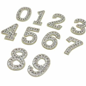 May include: Ten white numbers, 0 through 9, are covered in clear rhinestones. The numbers are arranged in a scattered pattern on a white background.