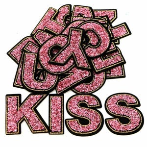 May include: Pink glitter iron-on letters spelling out the word "KISS" with black outlines. The letters are individually cut out and ready to be applied to fabric.