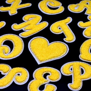 May include: A set of yellow felt letters and a heart shape, all outlined in white. The letters are in a variety of styles, including cursive and block letters.
