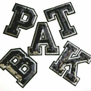 Peut inclure: Cinq lettres thermocollantes, P, A, T, B et K, sont en tissu noir avec des paillettes argent&eacute;es. Les lettres sont dans une police de caract&egrave;res de style universitaire.