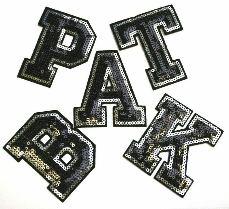 Peut inclure: Cinq lettres thermocollantes, P, A, T, B et K, sont en tissu noir avec des paillettes argent&eacute;es. Les lettres sont dans une police de caract&egrave;res de style universitaire.
