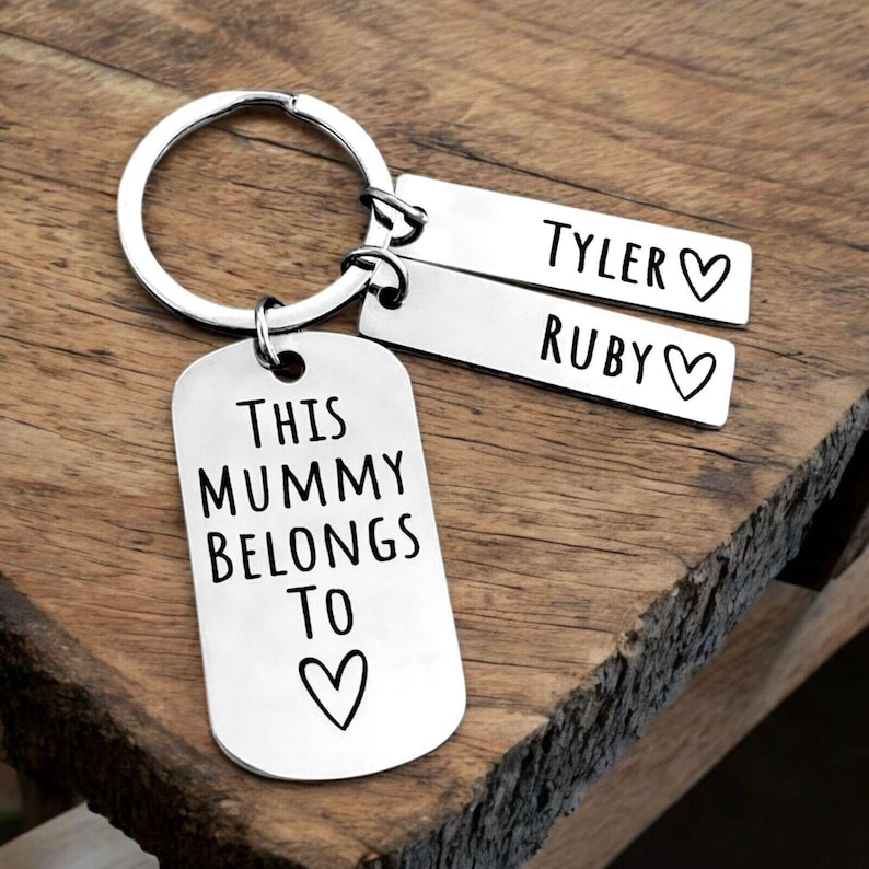 Puede incluir: Un llavero plateado con una etiqueta grande que dice "THIS MUMMY BELONGS TO" y un s&iacute;mbolo de coraz&oacute;n. Dos etiquetas m&aacute;s peque&ntilde;as est&aacute;n grabadas con los nombres "TYLER" y "RUBY" con s&iacute;mbolos de coraz&oacute;n. El llavero est&aacute; sobre una superficie de madera.
