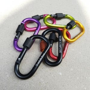 May include: A collection of six metal carabiners in various colors including black, purple, pink, lime green, yellow, and red. Each carabiner features a screw-lock gate mechanism and custom text engravings such as Grandads Keys and SECURITY KEYS.