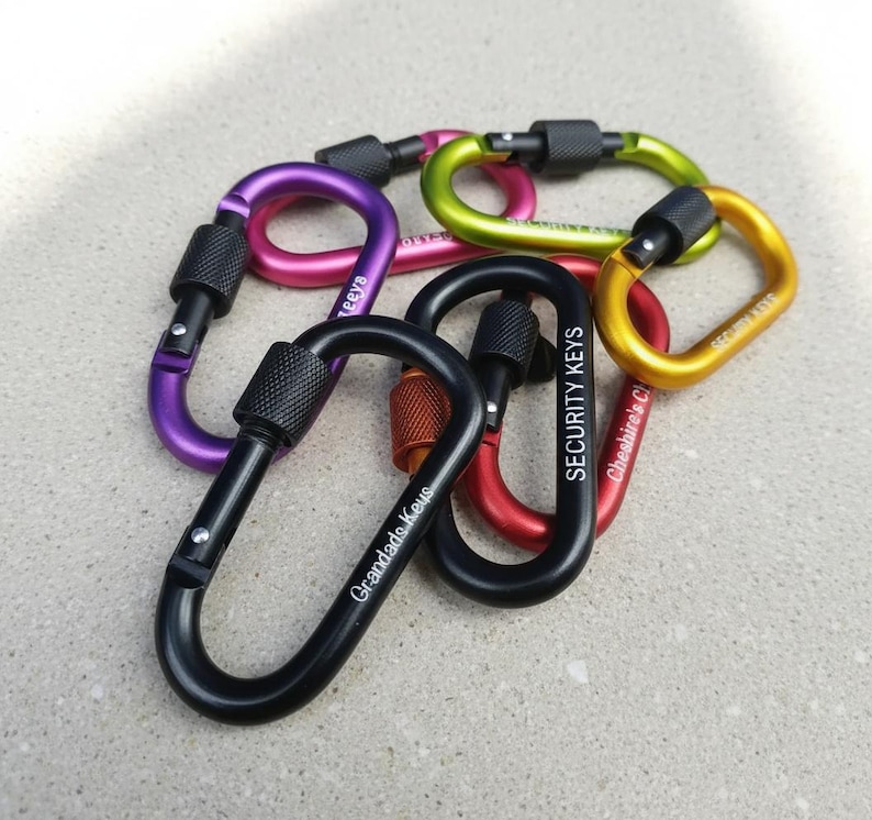 May include: A collection of six metal carabiners in various colors including black, purple, pink, lime green, yellow, and red. Each carabiner features a screw-lock gate mechanism and custom text engravings such as Grandads Keys and SECURITY KEYS.