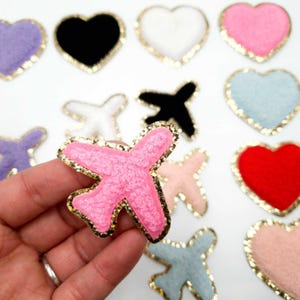 May include: A selection of colourful felt patches in the shape of aeroplanes and hearts. The pink aeroplane patch is held in a hand. The patches have gold glitter trim. Other patches are black, white, red, blue, and pink.