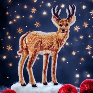 Pode incluir: Uma figura de veado bordada de cor castanho em pé sobre uma superfície branca e fofa com enfeites de Natal vermelhos em primeiro plano. O fundo é azul escuro com estrelas douradas.