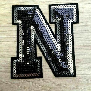 Peut inclure: Une lettre N en sequins noirs et argent&eacute;s. La lettre est faite de sequins cousus sur un support en tissu noir.