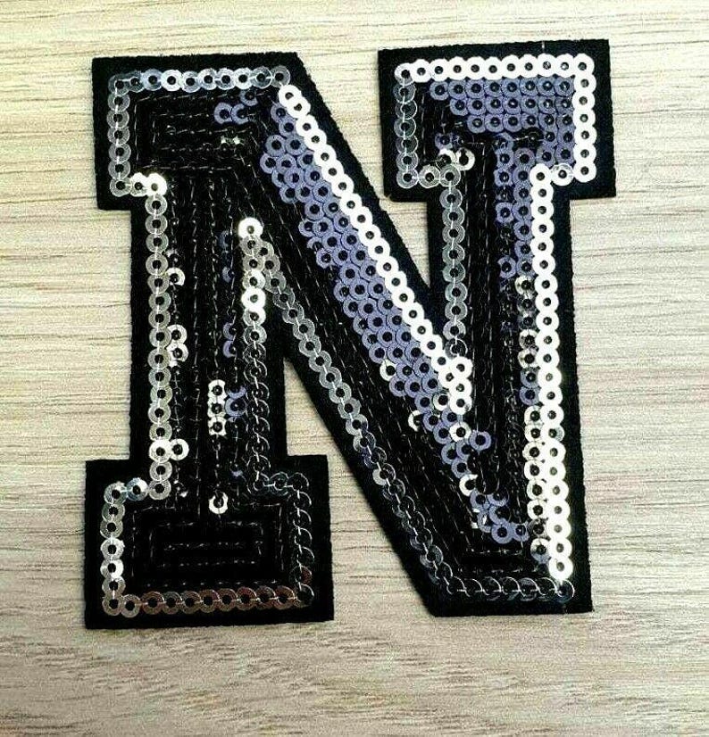 Peut inclure: Une lettre N en sequins noirs et argent&eacute;s. La lettre est faite de sequins cousus sur un support en tissu noir.