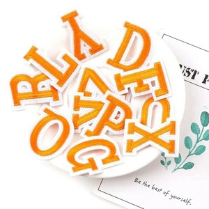 May include: A set of orange felt iron-on letters, including the letters A, B, D, E, F, O, Q, R, V, X, and Y. The letters are on a white surface.