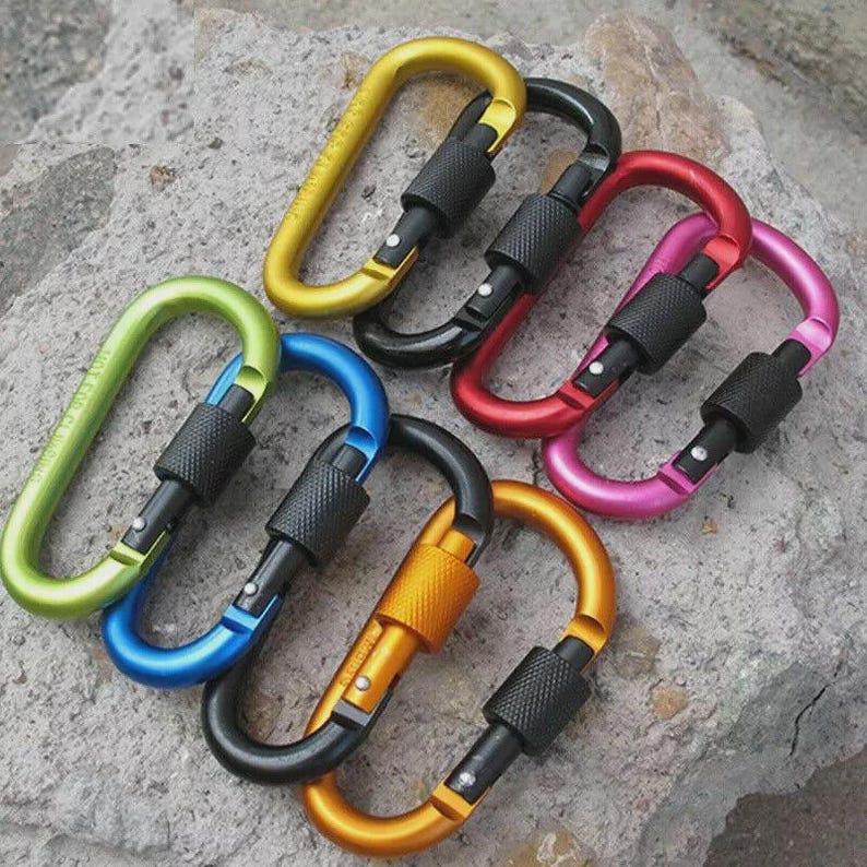 May include: A collection of six oval-shaped carabiners in various colors including yellow, black, red, pink, blue, and green. Each carabiner features a black metal screw-gate locking mechanism with a textured grip for secure closure and attachment.