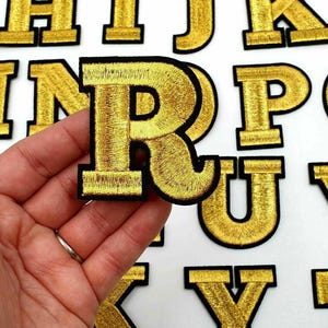 May include: Gold embroidered iron-on alphabet letters. The letters are in uppercase and are outlined in black. The letters are arranged in alphabetical order.