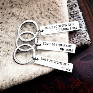 May include: Three silver keyrings with rectangular tags. Each tag reads "DON'T DO STUPID SHIT" followed by "MUM & DAD", "MUM", and "DAD", respectively. The tags also feature heart and floral designs. The keyrings are on a beige fabric.