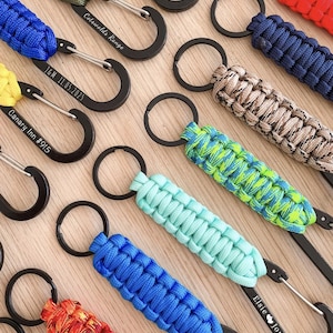 May include: A collection of paracord keychains featuring black metal carabiners with engraved text including West Lake Scouts, Cotswolds Range, J&M 11.05.2025, Canary Inn #915, and Elsie Jonah. The keychains display various braided patterns in colours like olive, blue, red, tan, green, and orange.
