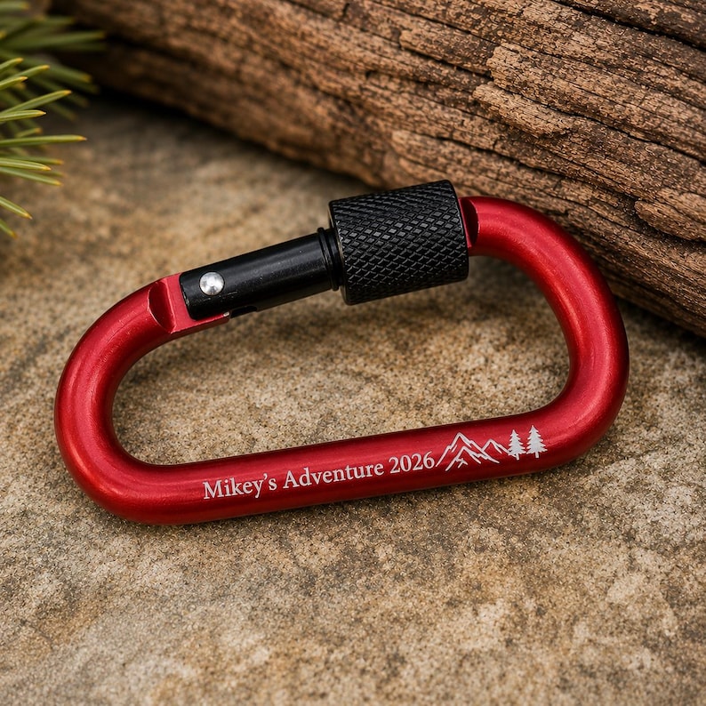 May include: A red metal carabiner with a black screw-lock gate. The carabiner features a white laser-engraved design consisting of the text Mikey's Adventure 2026 followed by a graphic illustration of mountain peaks and two pine trees.