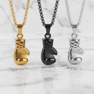 Puede incluir: Tres collares con colgantes de guantes de boxeo. Uno es dorado, otro negro y otro plateado. Cada colgante cuelga de una cadena a juego. Los colgantes est&aacute;n detallados con cordones y una forma redondeada.