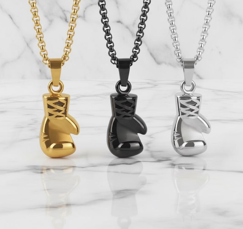 Puede incluir: Tres collares con colgantes de guantes de boxeo. Uno es dorado, otro negro y otro plateado. Cada colgante cuelga de una cadena a juego. Los colgantes est&aacute;n detallados con cordones y una forma redondeada.