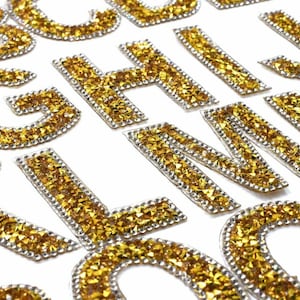 May include: A set of gold glitter iron-on letters, A through Z, with a silver outline. The letters are ideal for adding a touch of sparkle to clothing, bags, or other crafts.