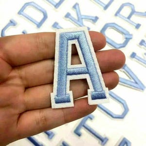 May include: A set of blue felt iron-on letters in various sizes. The letters are on a white background and have a white outline. The letter A is being held in a hand.