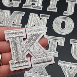 May include: Silver iron-on letters in the alphabet, including the letter K. The letters are on a white background.