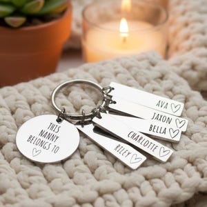 Puede incluir: Un llavero plateado con un dije redondo que dice "THIS NANNY BELONGS TO" y un coraz&oacute;n. Se adjuntan etiquetas rectangulares grabadas con nombres y s&iacute;mbolos de coraz&oacute;n. Los nombres incluyen Ava, Jaxon, Bella, Charlotte y Riley.