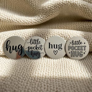 Könnte beinhalten: Vier silberfarbene, runde Token mit schwarzem Text. Die Token tragen die Wörter "hug", "little pocket hug", "hug" mit einem Herz und "little pocket hug" mit Sternen. Die Token liegen auf einer cremefarbenen, strukturierten Oberfläche.