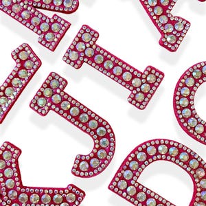 May include: Close-up of scattered, hot pink letters encrusted with iridescent rhinestones. The letters are in a variety of fonts and sizes, with a white background. The letters appear to be decorative, possibly for crafts or personalization.