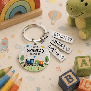 May include: A silver keychain with a round charm featuring a train illustration and the text "THIS GRANDAD BELONGS TO". Three tags are engraved with names: Ethan, Kyarra, and Amelia. Children's toys, including building blocks, surround the keychain.