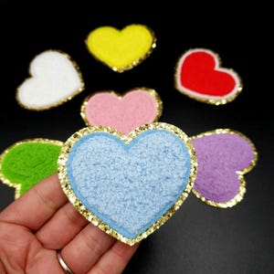 Puede incluir: Una colección de parches en forma de corazón de varios colores. Cada parche presenta un color diferente, incluyendo blanco, amarillo, rojo, rosa, verde, azul y morado. Los parches tienen un borde dorado brillante y una superficie texturizada. Los parches están sobre un fondo negro.