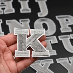 May include: A silver grey felt letter K with a white outline. The letter is held in a hand and there are other letters in the background.