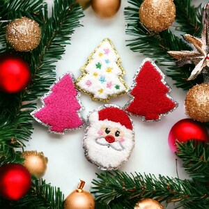 May include: A festive arrangement of Christmas ornaments and decorations. The image features four felt Christmas tree and Santa Claus ornaments in red, pink, and white, with gold and silver glitter trim. The scene is framed by green pine branches, red and gold baubles.