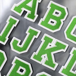 May include: Close-up of green embroidered varsity letter patches with white borders. The letters include A, B, I, J, and K. The patches are arranged on a grey surface.