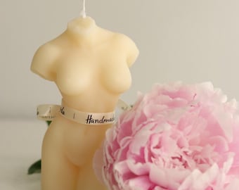 May include: A nude-colored candle shaped like a human torso with a white wick. The candle is wrapped with a white ribbon that says "Handmade". A pink peony flower is in the background.