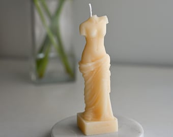 Op de afbeelding: Een witte kaars in de vorm van het standbeeld van de Venus van Milo op een witte marmeren voet. De kaars staat voor een vaas met gele calla's.