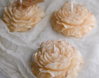 May include: Three ivory-colored rose-shaped candles with a gold shimmer. The candles are on a white fabric background. One candle has a small brown tag with the word "remarkable" written on it.
