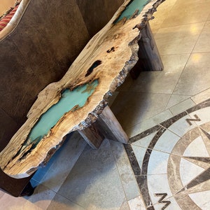 May include: A live edge wooden table with a turquoise resin river running through the center. The table is supported by two wooden legs. The wood has a natural, light brown color with dark knots and a rough edge.