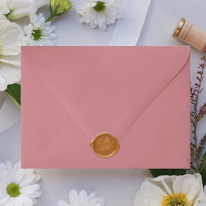 May include: A blush pink envelope with a gold wax seal. The seal has a floral design. The envelope is surrounded by white flowers.