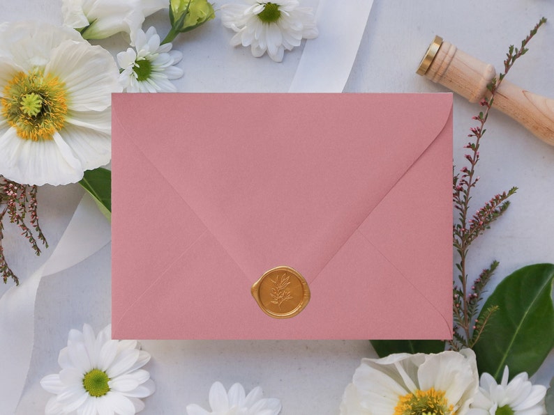 May include: A blush pink envelope with a gold wax seal. The seal has a floral design. The envelope is surrounded by white flowers.