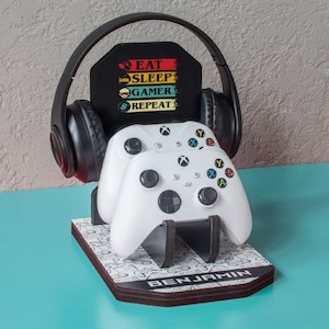 Può includere: Un supporto per cuffie da gioco bianco e nero con cuffie nere, due controller Xbox bianchi e un cartello che dice "Mangia Dormi Gioca Ripeti" con un'icona di giocatore. Il supporto è personalizzato con il nome "Benjamin".