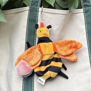 Könnte beinhalten: Ein Plüsch-Bienen-Schlüsselanhänger mit gelb-schwarz gestreiftem Körper, orangefarbenen, strukturierten Flügeln und einem kleinen silbernen Metallclip. Ein herzförmiger Kunststoff-Etikettenhalter ist an einem Flügel befestigt, ein weißes Etikett hängt.