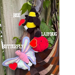 K&ouml;nnte beinhalten: Drei Pl&uuml;sch-Schl&uuml;sselanh&auml;nger in Form von Insekten und Tieren an einer braunen Ledertasche. Die Artikel umfassen eine gelb-schwarze Biene, einen roten Marienk&auml;fer mit schwarzen Punkten und einen pastellfarbenen rosa-blauen Schmetterling mit Herz.